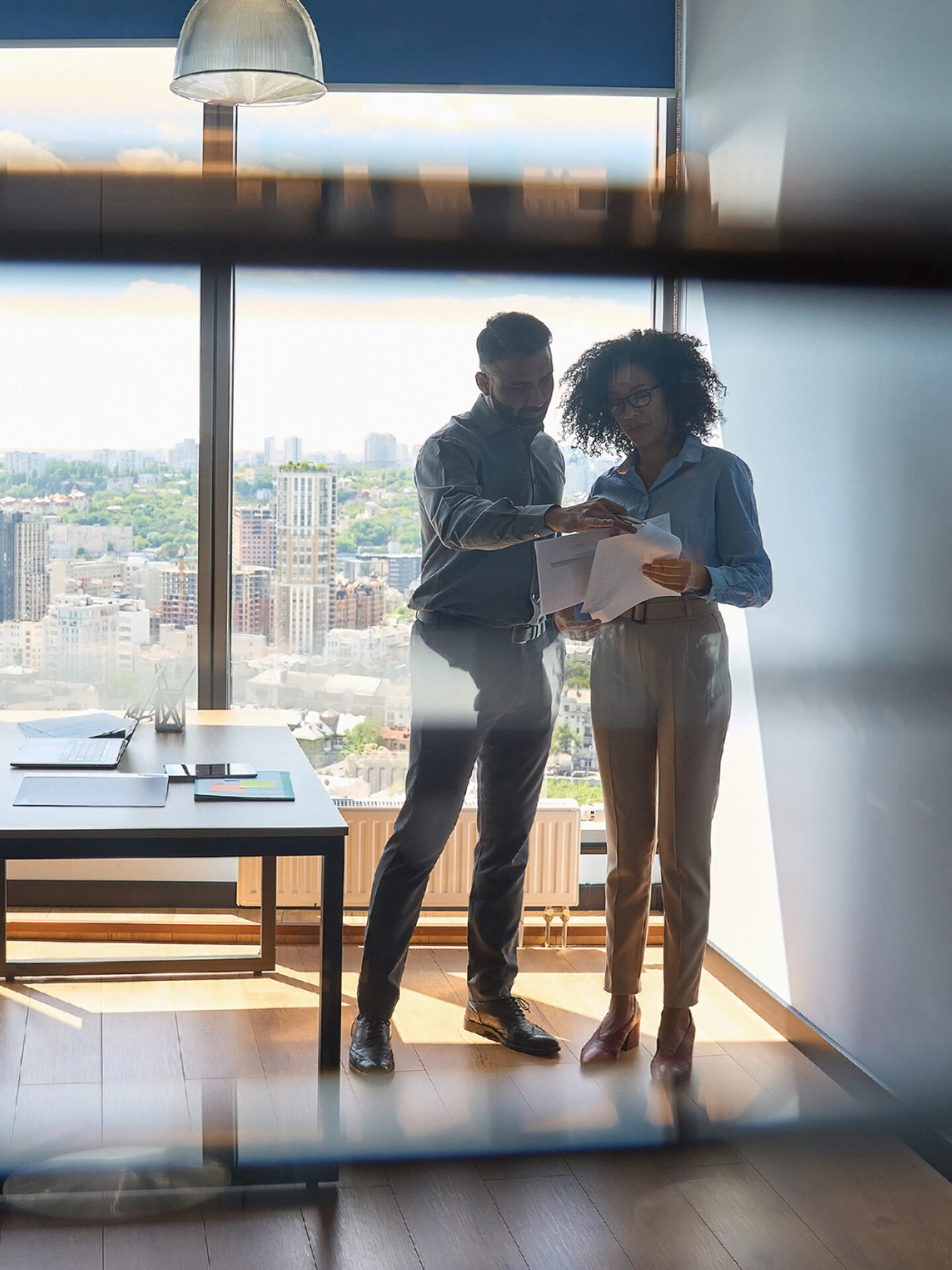 Multiracial colleagues standing discussing report paperwork in modern office. Indian ceo advisor male boss mentor explaining financial analytics report to African American female intern holding paper documents standing in modern office. Shot through jalousie glass.